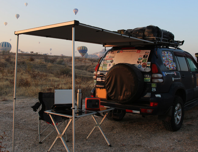 Coche 4x4 con un toldo trasero y una mesa de camping. En el fonto, un panorama al atardecer con globos aerostáticos.